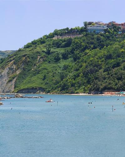 A coastal scene showing a sandy beach crowded with people, turquoise water, and a steep, green hillside with buildings, including a prominent white hotel, rising above the shore.