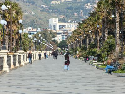 A wide, paved waterfront promenade lined with tall palm trees and distinctive spherical streetlights on black posts, with people walking and sitting on benches, overlooking a hillside town and sea in the distance.