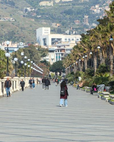 A wide, paved waterfront promenade lined with tall palm trees and distinctive spherical streetlights on black posts, with people walking and sitting on benches, overlooking a hillside town and sea in the distance.