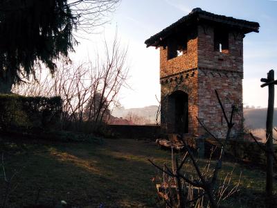 A weathered brick tower with an arched ground-floor opening and open belfry-like openings on the upper level, set in a grassy, wooded landscape with bare trees and a wooden cross nearby.