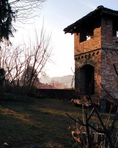 A weathered brick tower with an arched ground-floor opening and open belfry-like openings on the upper level, set in a grassy, wooded landscape with bare trees and a wooden cross nearby.