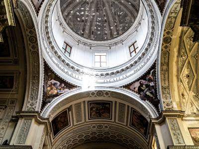 An interior upward view of a large, ornate dome with a central oculus, surrounded by frescoes, Latin inscriptions, and decorative architectural elements within a church.