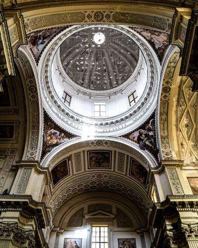 An interior upward view of a large, ornate dome with a central oculus, surrounded by frescoes, Latin inscriptions, and decorative architectural elements within a church.