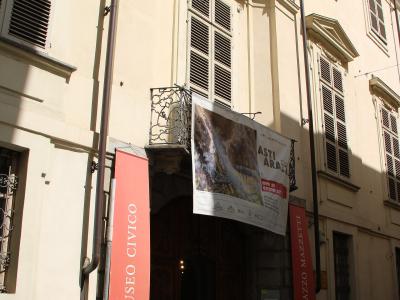 The image shows the exterior facade of a historic building with a large arched entrance, flanked by vertical banners reading "MUSEO CIVICO" and "PALAZZO MAZZETTI," and a banner advertising an exhibition titled "ASTI ARAZZI."