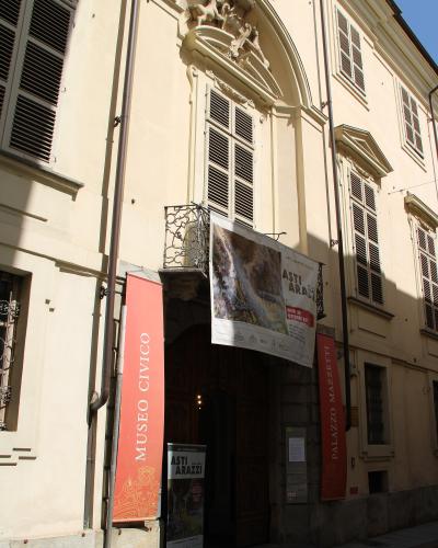 The image shows the exterior facade of a historic building with a large arched entrance, flanked by vertical banners reading "MUSEO CIVICO" and "PALAZZO MAZZETTI," and a banner advertising an exhibition titled "ASTI ARAZZI."