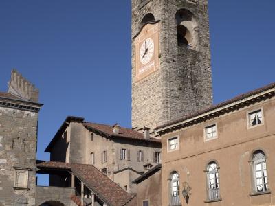 A tall medieval stone bell tower with a large clock face, situated in a paved public square surrounded by historic buildings and outdoor café seating under a clear blue sky.