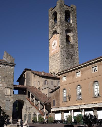 A tall medieval stone bell tower with a large clock face, situated in a paved public square surrounded by historic buildings and outdoor café seating under a clear blue sky.