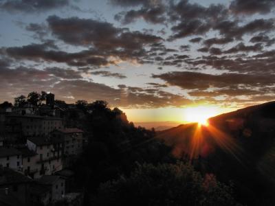 A sunset view over a hillside village with terracotta-roofed buildings and a prominent tower, set against a dramatic cloudy sky with sunburst rays.