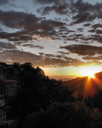 A sunset view over a hillside village with terracotta-roofed buildings and a prominent tower, set against a dramatic cloudy sky with sunburst rays.