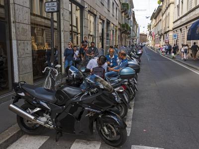A row of parked motorcycles and scooters along a city street curb, with pedestrians walking on the sidewalk and storefronts visible in the background.