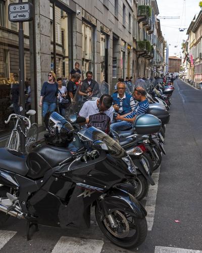 A row of parked motorcycles and scooters along a city street curb, with pedestrians walking on the sidewalk and storefronts visible in the background.