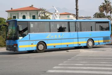A light blue Setra intercity coach with "Lazzi" written in yellow script on the side, parked on a paved street with Italian-style buildings, palm trees, and a modern sculpture in the background.