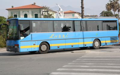 A light blue Setra intercity coach with "Lazzi" written in yellow script on the side, parked on a paved street with Italian-style buildings, palm trees, and a modern sculpture in the background.