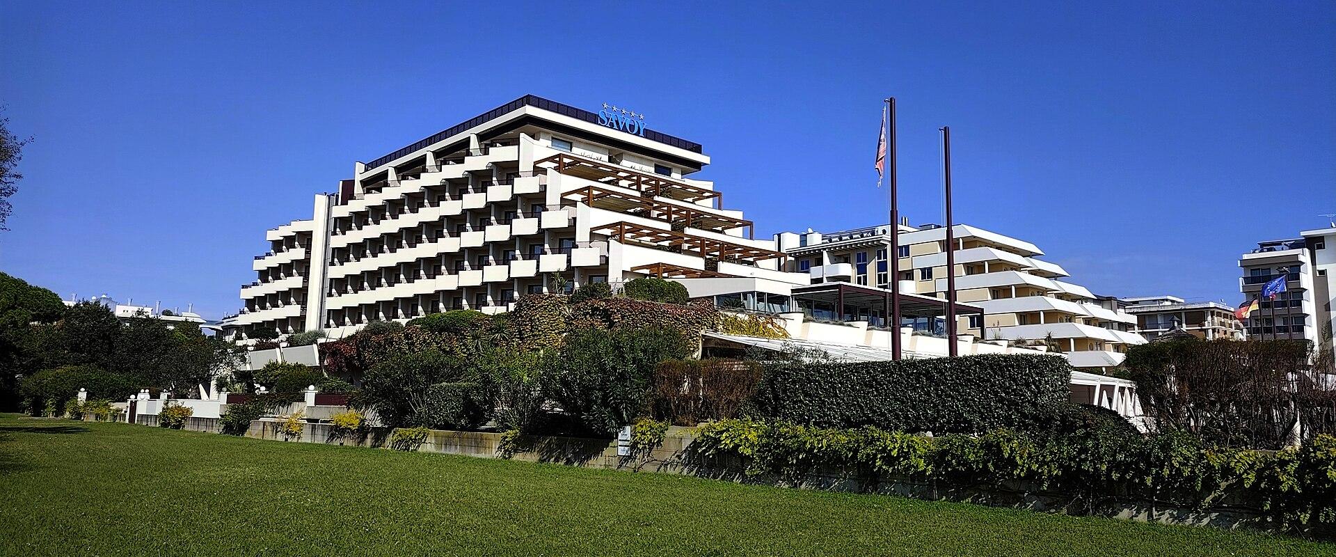 A large, modern white hotel building with tiered balconies and a wooden pergola structure on the roof, set against a clear blue sky, with green lawns and shrubbery in the foreground; signage on the building reads “SAVOY”.