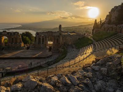 A wide-angle photograph of an ancient stone amphitheater with tiered seating, a stage area, and ruins on a hillside, overlooking the sea and distant mountains during sunset.