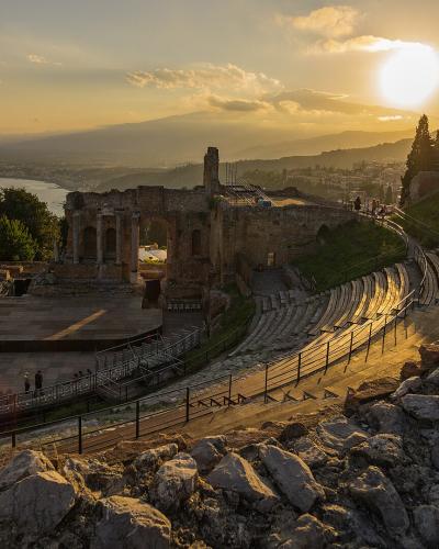 A wide-angle photograph of an ancient stone amphitheater with tiered seating, a stage area, and ruins on a hillside, overlooking the sea and distant mountains during sunset.