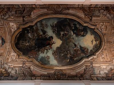 An interior ceiling fresco featuring a central oval painting of religious figures amidst clouds, surrounded by ornate trompe-l'œil architectural stucco work and two circular medallions containing black birds.