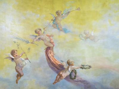 The image shows a classical-style fresco painting featuring several cherubs (putti) in flight against a soft yellow and blue sky, holding musical instruments, laurel wreaths, and ribbons.