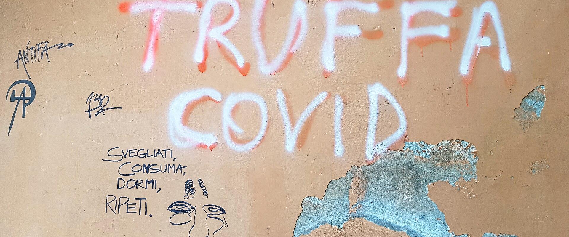 A close-up of a weathered beige wall covered in graffiti, prominently featuring the words "TRUFFA COVID" in large white spray paint, along with smaller handwritten text including "SVEGLIATI, CONSUMA, DORMI, RIPETI." and a simple line drawing of a