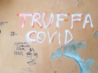 A close-up of a weathered beige wall covered in graffiti, prominently featuring the words "TRUFFA COVID" in large white spray paint, along with smaller handwritten text including "SVEGLIATI, CONSUMA, DORMI, RIPETI." and a simple line drawing of a