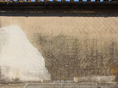 A close-up of a weathered, textured exterior wall featuring faint geometric and floral stucco patterns, with a large patch of lighter plaster on the left and dark staining on the right, topped by a tiled roof edge.