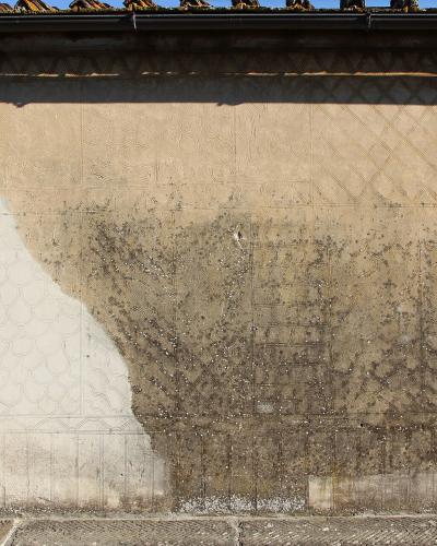 A close-up of a weathered, textured exterior wall featuring faint geometric and floral stucco patterns, with a large patch of lighter plaster on the left and dark staining on the right, topped by a tiled roof edge.