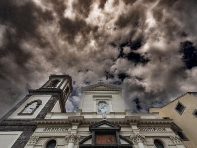 A low-angle photograph of a white church facade with a dark stone bell tower on the left, featuring the inscriptions "BASILICH" and "PONTIFICIA" on the entablature, set against a dramatic cloudy sky.