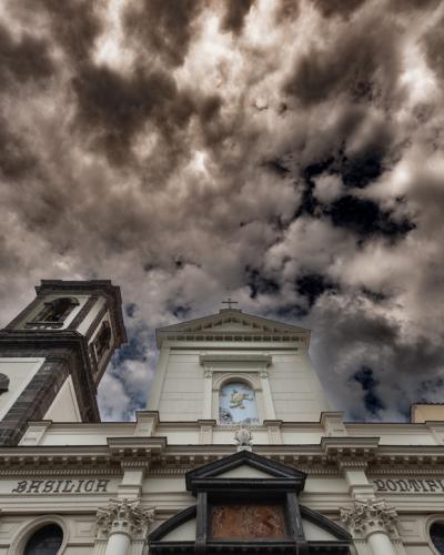 A low-angle photograph of a white church facade with a dark stone bell tower on the left, featuring the inscriptions "BASILICH" and "PONTIFICIA" on the entablature, set against a dramatic cloudy sky.