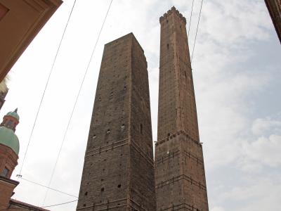 A low-angle view of two tall, medieval brick towers standing side-by-side against a cloudy sky, with a portion of a building featuring a green dome visible on the left and overhead wires crossing the frame.