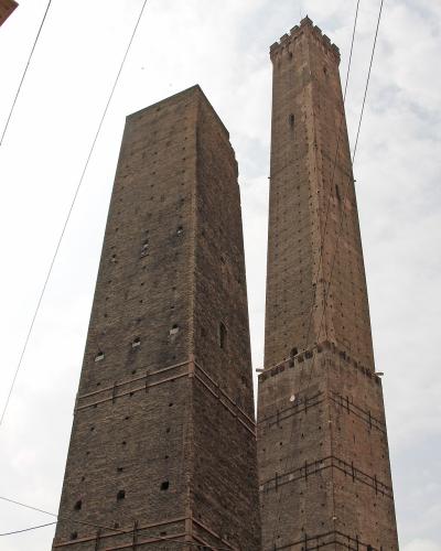 A low-angle view of two tall, medieval brick towers standing side-by-side against a cloudy sky, with a portion of a building featuring a green dome visible on the left and overhead wires crossing the frame.