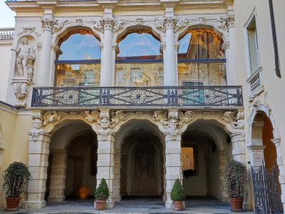 The image shows the interior courtyard (corte) of a Baroque palace featuring a two-story loggia with ornate stone columns, sculpted masks, and a decorative wrought-iron balcony, set above a geometric pebble mosaic pavement.