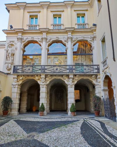 The image shows the interior courtyard (corte) of a Baroque palace featuring a two-story loggia with ornate stone columns, sculpted masks, and a decorative wrought-iron balcony, set above a geometric pebble mosaic pavement.