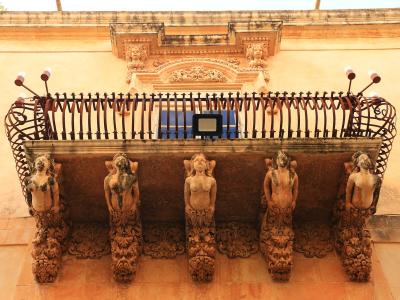A close-up view of an ornate stone balcony supported by five sculpted female figures (caryatids) with elaborate iron railings and decorative stonework above, set against a warm-toned building facade.