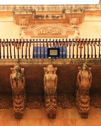 A close-up view of an ornate stone balcony supported by five sculpted female figures (caryatids) with elaborate iron railings and decorative stonework above, set against a warm-toned building facade.