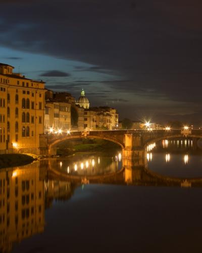 A night-time photograph of the Ponte Santa Trinita bridge spanning the Arno River in Florence, with illuminated historic buildings and a bell tower on the left bank (Oltrarno) and their reflections in the water.