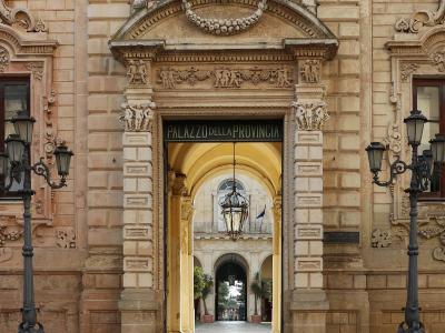A Baroque stone archway entrance with ornate carvings, flanked by black lampposts, leading into a courtyard; signage above the arch reads "PALAZZO DELLA PROVINCIA".