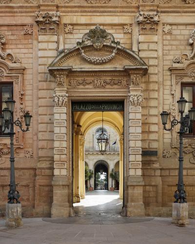 A Baroque stone archway entrance with ornate carvings, flanked by black lampposts, leading into a courtyard; signage above the arch reads "PALAZZO DELLA PROVINCIA".