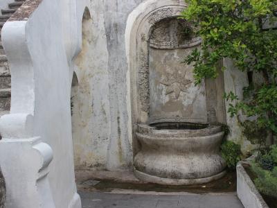 A weathered stone fountain with a star-shaped relief set into an arched niche, flanked by a white plastered staircase on the left and green foliage on the right, within a terraced garden setting.