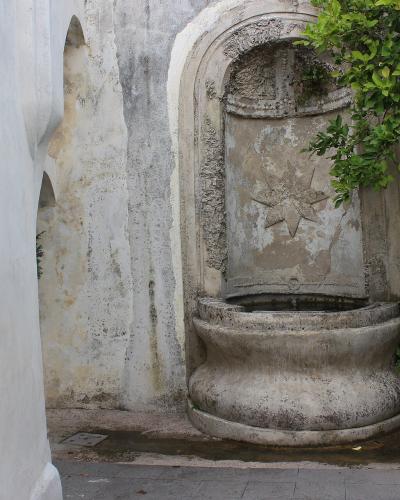 A weathered stone fountain with a star-shaped relief set into an arched niche, flanked by a white plastered staircase on the left and green foliage on the right, within a terraced garden setting.