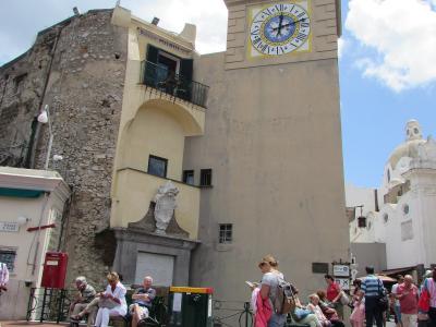 The image shows a sunlit public square with a prominent clock tower, a stone building with a balcony, a red postbox, benches with people sitting, and a white church dome in the background; signage includes “Ristorante Pulacchi” and “PIAZZA A PIAZ”