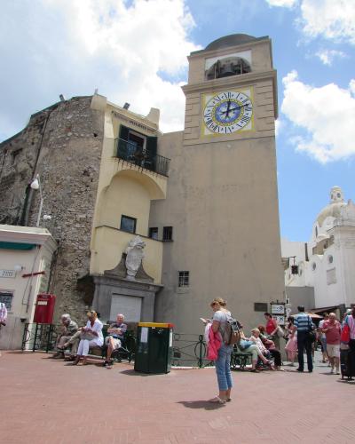 The image shows a sunlit public square with a prominent clock tower, a stone building with a balcony, a red postbox, benches with people sitting, and a white church dome in the background; signage includes “Ristorante Pulacchi” and “PIAZZA A PIAZ”