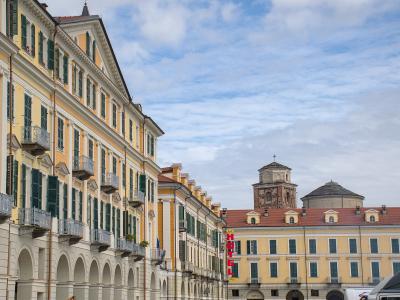 A street-level view of a wide, arcaded Italian piazza with yellow and cream buildings, green shutters, balconies, and a distant church tower under a cloudy sky; a red vertical "HOTEL" sign is visible on the right side.