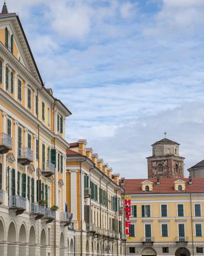 A street-level view of a wide, arcaded Italian piazza with yellow and cream buildings, green shutters, balconies, and a distant church tower under a cloudy sky; a red vertical "HOTEL" sign is visible on the right side.