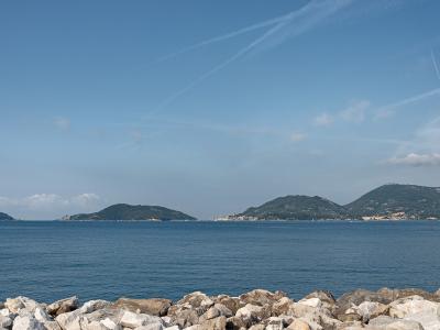 A coastal seascape showing a rocky foreground, calm blue water, and distant green hills/islands under a sky with contrails; no visible signage or specific landmarks are clearly identifiable.