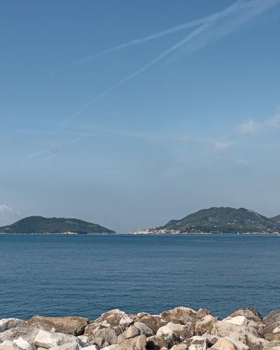 A coastal seascape showing a rocky foreground, calm blue water, and distant green hills/islands under a sky with contrails; no visible signage or specific landmarks are clearly identifiable.