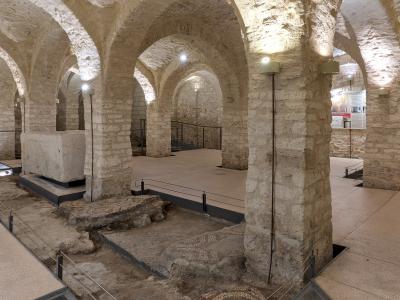 The image shows the interior of a stone crypt or underground archaeological area with vaulted ceilings, thick pillars, exposed ancient mosaic floors, and modern lighting and railings for visitors.