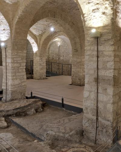 The image shows the interior of a stone crypt or underground archaeological area with vaulted ceilings, thick pillars, exposed ancient mosaic floors, and modern lighting and railings for visitors.
