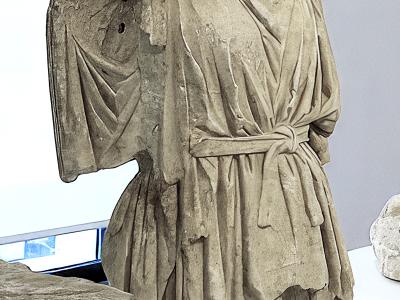 A headless ancient marble statue of a draped figure, likely from antiquity, displayed on metal supports in a modern museum gallery with white walls and a window visible in the background.