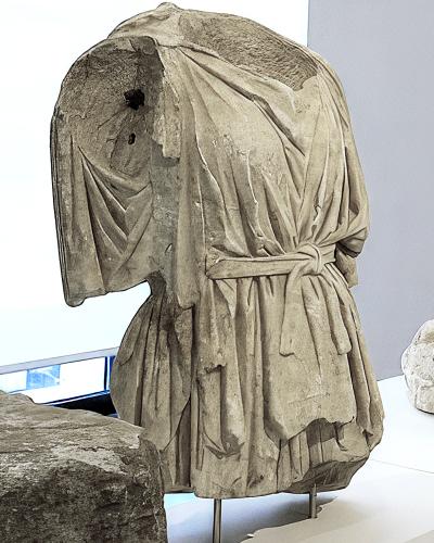 A headless ancient marble statue of a draped figure, likely from antiquity, displayed on metal supports in a modern museum gallery with white walls and a window visible in the background.