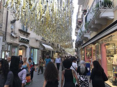 A crowded pedestrian street in an Italian town, lined with shops like "GEOX" and "RUSSOTTI," decorated with hanging gold and silver streamers.